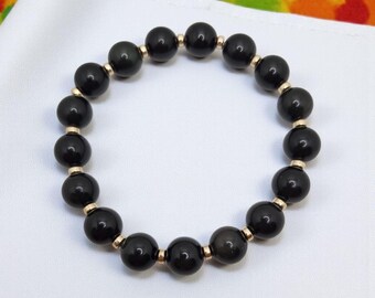 May include: A black beaded bracelet with gold-coloured spacers. The bracelet features round, polished black beads strung together with small gold-coloured beads between each black bead. It is displayed on a white surface.