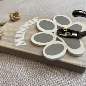 May include: A wooden dog leash hook with a grey paw print design and a black metal hook. The hook is labelled "MINNIE".