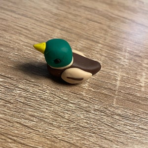 Puede incluir: Una pequeña figurita de pato de arcilla hecha a mano. El pato está pintado de verde, marrón y amarillo.