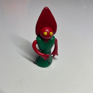 May include: A red and green clay figurine with a pointed head and yellow eyes. The figurine is wearing a green robe with a black belt.