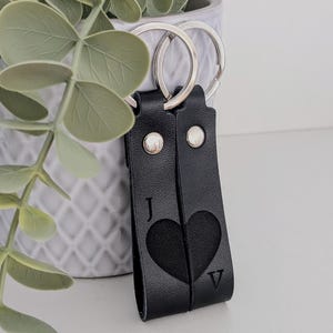 Könnte beinhalten: Schwarzer Leder-Schlüsselanhänger mit einem Herz-Ausschnitt und den Buchstaben "J" und "V". Der Schlüsselanhänger hat zwei silberne Schlüsselringe und ist genietet. Eine dekorative Pflanze in einem grauen Topf befindet sich im Hintergrund.