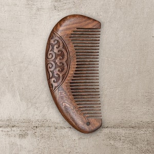 May include: A dark brown wooden comb with a carved design on the back. The comb is shaped like a crescent moon and has fine teeth.
