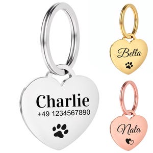 Op de afbeelding: Hartvormige huisdier-ID-tags in zilver, goud en roségoud. De zilveren tag heeft "Charlie" en een telefoonnummer, met een pootafdruk. De gouden tag zegt "Bella" met een pootafdruk. De roségouden tag zegt "Nala" met een hartje.