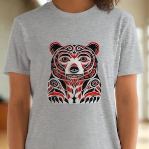 May include: A light gray t-shirt featuring a detailed bear design in red, black, and white. The bear graphic is centered on the shirt, showcasing intricate patterns and shapes. The shirt is made of a soft material.