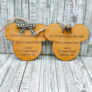 May include: Two wooden ornaments shaped like Mickey Mouse ears with a black and white checkered ribbon and a silver bell. The ornaments have the text "You've been pixie dusted! Have a magical day! Love, The Kunz Family Jackson, WI" on them.