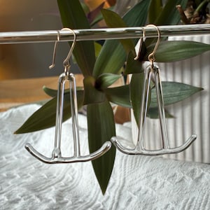 May include: A pair of silver-colored earrings with a unique, geometric design. The earrings have a hook closure and a curved, horizontal bar at the bottom. The earrings are displayed against a backdrop of green leaves and a white, textured surface.