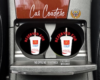 May include: Black car coasters with a McDonald's drink design. The coasters are black with the words "RESERVED FOR" in red around a white cup with the McDonald's logo and Coca-Cola branding. The text "Car Coasters" is above the coasters.
