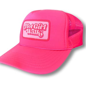 K&ouml;nnte beinhalten: Eine rosafarbene Trucker-Kappe mit wei&szlig;em Netzr&uuml;cken. Die Kappe hat einen wei&szlig;en Aufn&auml;her mit dem Text "Hot Girl Walk" in rosa Buchstaben.