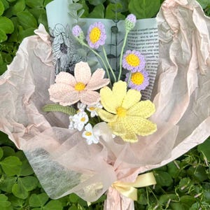 May include: A bouquet of handmade crochet flowers in shades of pink, yellow, and purple, wrapped in light pink tissue paper and tied with a yellow ribbon. The bouquet includes daisies and other flower types, creating a charming floral arrangement.