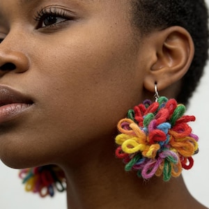 May include: Close-up of a person wearing large, colorful, handmade earrings. The earrings are made of looped yarn in shades of red, yellow, blue, green, and purple. The earrings are attached to silver hooks.