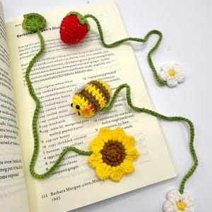 May include: Crocheted bookmarks featuring a red strawberry, a yellow and brown bee, a yellow sunflower, and white daisies. The bookmarks are connected by green stems and placed on an open book.