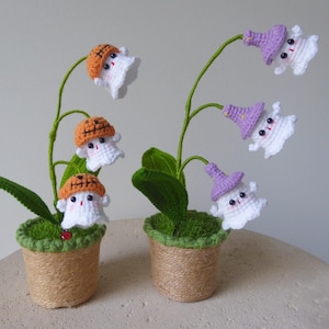 May include: Two potted plants with white crocheted flowers shaped like ghosts. The flowers have different colored hats, including orange, purple, and a pumpkin-shaped hat. The plants are in brown pots with green moss and twine.