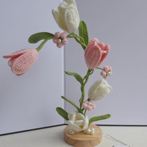 Może przedstawiać: Lampka z kwiatów wykonana na szydełku z różowymi i białymi tulipanami, zielonymi liśćmi i perłowymi akcentami. Lampka stoi na drewnianej podstawie i jest wyposażona w pilot zdalnego sterowania i zasilacz.