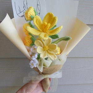 May include: A handmade bouquet of yellow and white crochet flowers, wrapped in beige paper and burlap. The bouquet includes a variety of flower types, with green stems and leaves. A white ribbon with text is tied around the base.
