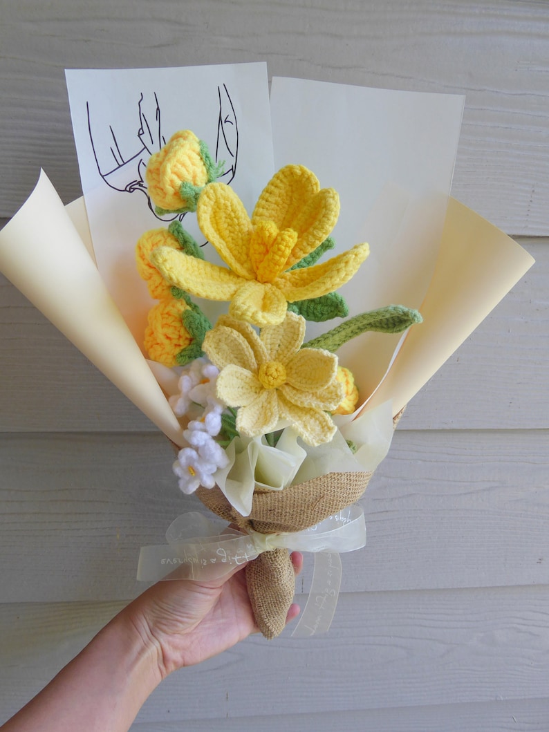 May include: A handmade bouquet of yellow and white crochet flowers, wrapped in beige paper and burlap. The bouquet includes a variety of flower types, with green stems and leaves. A white ribbon with text is tied around the base.