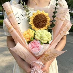May include: A bouquet of crocheted flowers, including a sunflower, roses, and other blooms, wrapped in pink paper with text. The bouquet is tied with a pink ribbon. The word "LOVE" is visible on the wrapping.