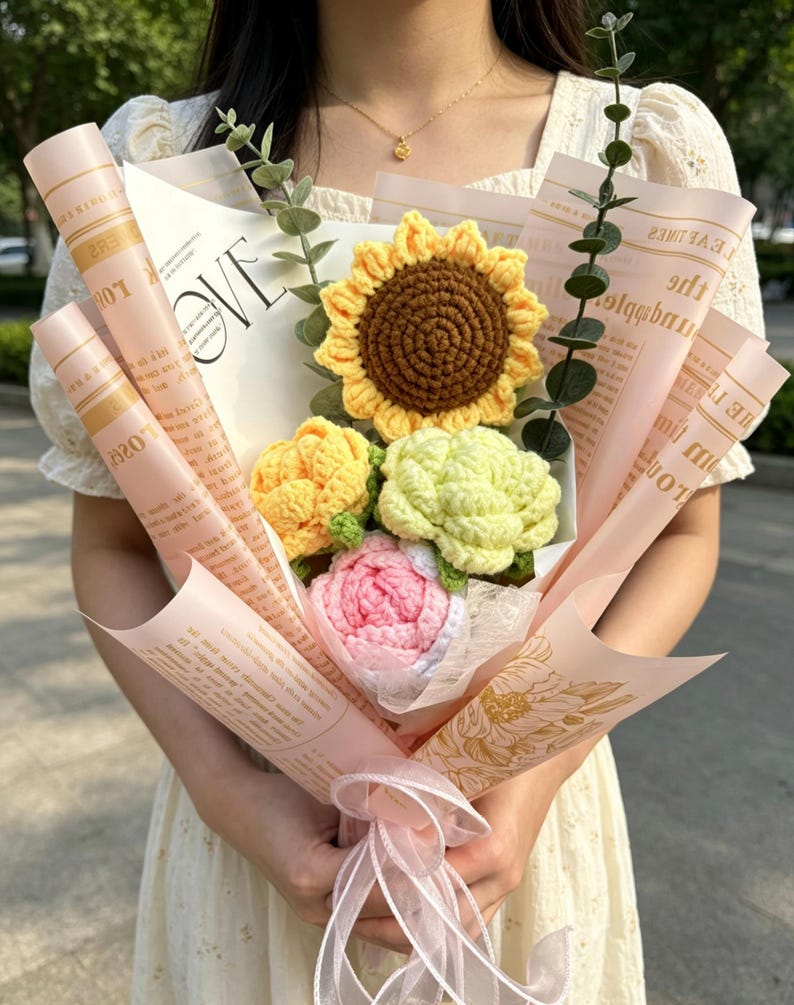 May include: A bouquet of crocheted flowers, including a sunflower, roses, and other blooms, wrapped in pink paper with text. The bouquet is tied with a pink ribbon. The word "LOVE" is visible on the wrapping.