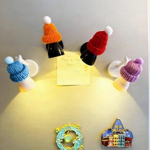 May include: A collection of refrigerator magnets, including four small lamps with knitted hat shades in orange, red, blue, and purple. A yellow sticky note with the text "Have a Nice Day!" is also attached. Additional magnets include a New Orleans souvenir and a teal and gold circular design.