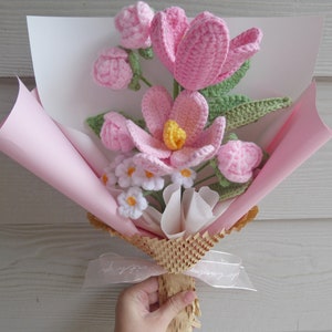 May include: A handmade crochet bouquet featuring pink flowers with yellow centres and green leaves. The bouquet is wrapped in pink and white paper, tied with a ribbon, and held by a hand.