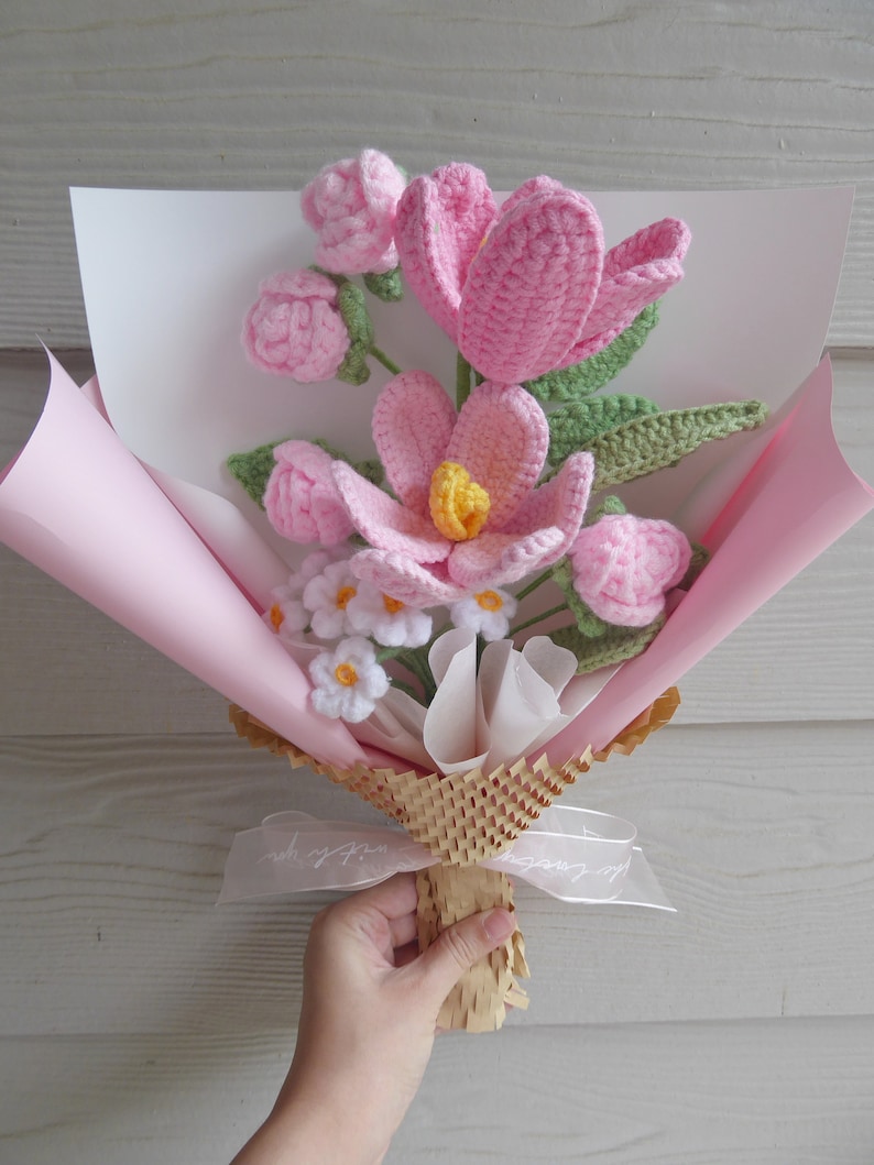 May include: A handmade crochet bouquet featuring pink flowers with yellow centres and green leaves. The bouquet is wrapped in pink and white paper, tied with a ribbon, and held by a hand.