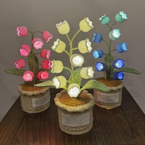 May include: Three crochet flower lamps in pots. The lamps feature pink, yellow, and blue flowers with glowing lights. The pots are tan and have a woven texture, sitting on a wooden surface.