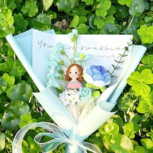 May include: A bouquet of crocheted flowers in shades of blue, white, and green, wrapped in light blue paper and tied with a sheer ribbon. A card with the words "You are sunshine" is included. The bouquet features a crocheted doll.