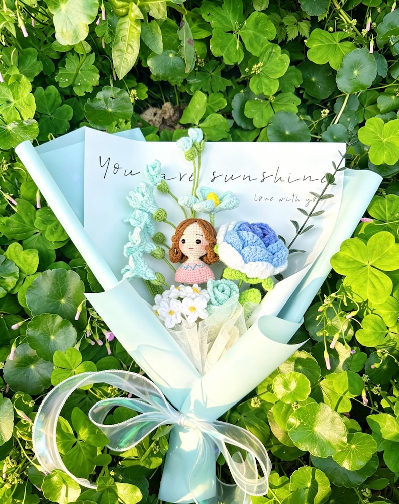 May include: A bouquet of crocheted flowers in shades of blue, white, and green, wrapped in light blue paper and tied with a sheer ribbon. A card with the words "You are sunshine" is included. The bouquet features a crocheted doll.