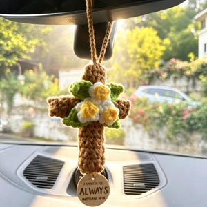 May include: A brown crocheted cross hanging ornament featuring a cluster of white and yellow crocheted flowers with green leaves. A circular wooden tag hangs from the bottom, engraved with the text I AM WITH YOU ALWAYS MATTHEW 28:20.