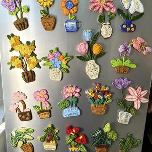 以下が含まれることがあります： 様々なスタイルの、カラフルなかぎ針編みの花のマグネットのコレクション。ひまわり、チューリップ、蘭など、それぞれが装飾的な鉢や花瓶に入っています。カラーパレットは、黄色、ピンク、青、白で、茶色の鉢と緑の葉があります。