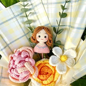 May include: A crochet bouquet with a doll and various flowers. The doll has brown hair and a pink dress. The flowers include a pink rose, a yellow and orange rose, and a white daisy. The bouquet is wrapped in yellow and white check paper.