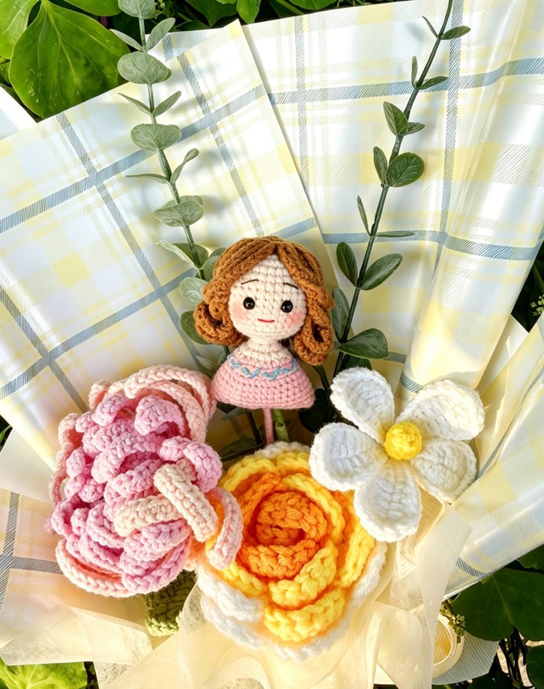 May include: A crochet bouquet with a doll and various flowers. The doll has brown hair and a pink dress. The flowers include a pink rose, a yellow and orange rose, and a white daisy. The bouquet is wrapped in yellow and white check paper.