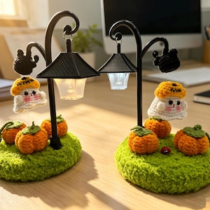 Op de afbeelding: Twee zwarte metalen straatlantaarns met hangende gehaakte spookjes met oranje pompoenmutsen. Elke lamp staat op een groene gehaakte basis versierd met kleine oranje pompoenen en een piepkleine zwarte spin.