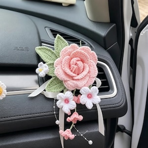 May include: A handmade crochet car decoration featuring a large pink rose, green leaves, and small white flowers. The floral arrangement includes white ribbons and is attached to a car dashboard. The accessory is a decorative item.