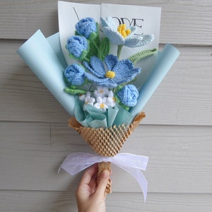 May include: A handcrafted crochet flower bouquet featuring blue, white, and yellow blooms, presented in light blue paper and tied with a white ribbon. The bouquet includes different flower styles and green leaves. A card with the word "LOVE" is visible.