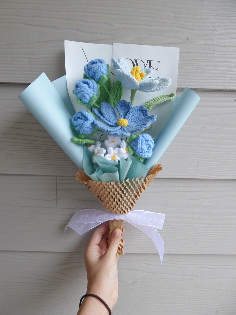 May include: A handcrafted crochet flower bouquet featuring blue, white, and yellow blooms, presented in light blue paper and tied with a white ribbon. The bouquet includes different flower styles and green leaves. A card with the word "LOVE" is visible.