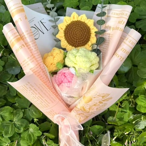 May include: A handmade bouquet of crocheted flowers, featuring a sunflower, a peony, and other blooms, wrapped in pink paper with printed text. The bouquet is tied with a sheer ribbon, set against a backdrop of green foliage.