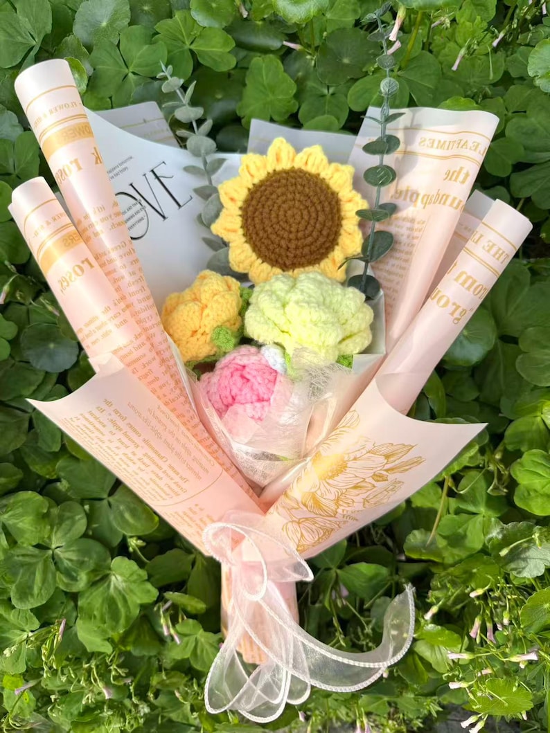 May include: A handmade bouquet of crocheted flowers, featuring a sunflower, a peony, and other blooms, wrapped in pink paper with printed text. The bouquet is tied with a sheer ribbon, set against a backdrop of green foliage.