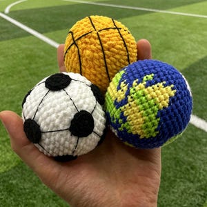 May include: Three crocheted balls: a yellow basketball, a soccer ball with black and white panels, and a blue and green globe. The balls are held in a hand against a green field background.