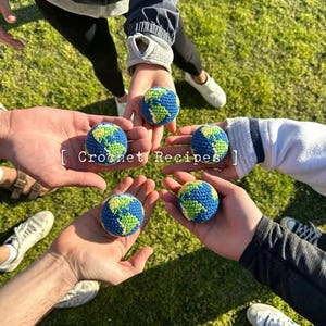 May include: Several hands holding crocheted Earth globes. The globes are blue with green and yellow continents. The text "Crochet Recipes" is visible in the image. The background is green grass.