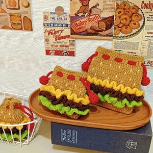May include: Crocheted burger-shaped pouches in shades of brown, yellow, and green, with red drawstring closures. The pouches are displayed on a wooden tray and in a white wire basket. Vintage-style advertisements are in the background.