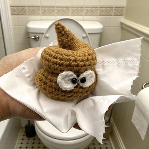 May include: A brown crocheted poop emoji with white eyes and black pupils, on white toilet paper. The background shows a toilet and bathroom fittings.