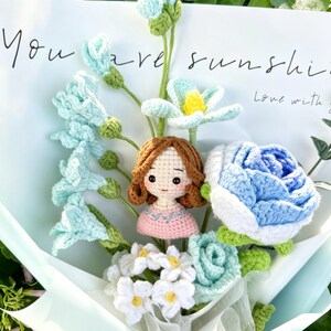 May include: A handcrafted crochet bouquet with blue and white flowers, a small doll, and the words "You are sunshine." The bouquet is wrapped in light blue paper, creating a unique floral arrangement.