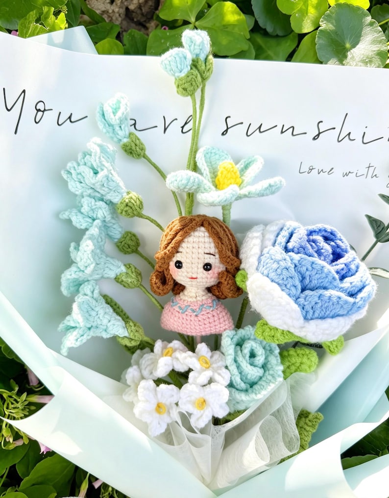 May include: A handcrafted crochet bouquet with blue and white flowers, a small doll, and the words "You are sunshine." The bouquet is wrapped in light blue paper, creating a unique floral arrangement.