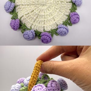 May include: A round, cream-colored crocheted doily featuring a border of green leaves and small, light purple crocheted roses. Below, a small, mustard-yellow crocheted basket with a handle is filled with light purple crocheted roses and green leaves.