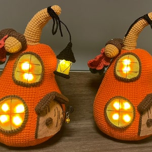 Puede incluir: Dos casas de punto de crochet con forma de calabaza con ventanas iluminadas y una pequeña linterna de crochet colgando de la parte superior. Las casas son de color naranja y marrón con una puerta marrón y una pequeña campana colgando de la puerta.