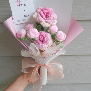 May include: A bouquet of crocheted pink roses and buds, wrapped in pink paper and tied with a peach ribbon. A card with the words "FALL in love" is attached with a pink paper clip. The bouquet is held by a person.