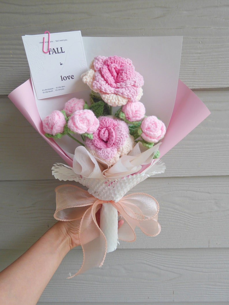 May include: A bouquet of crocheted pink roses and buds, wrapped in pink paper and tied with a peach ribbon. A card with the words "FALL in love" is attached with a pink paper clip. The bouquet is held by a person.