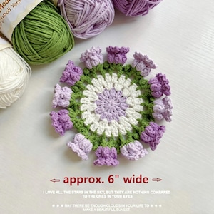 May include: A round crochet doily featuring a white center, a light purple inner ring, and a green outer border adorned with twelve small, three-dimensional purple tulip-shaped flowers. The text below states approx. 6" wide.