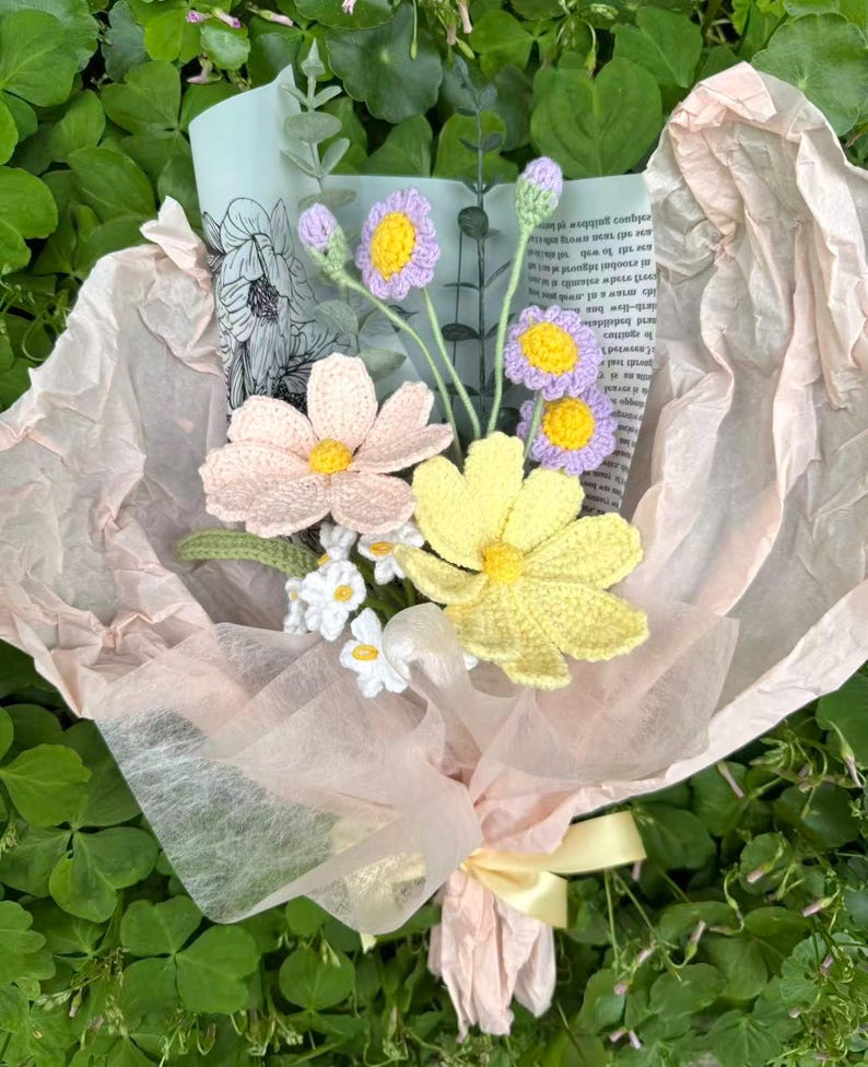 May include: A bouquet of handmade crochet flowers in shades of pink, yellow, and purple, wrapped in light pink tissue paper and tied with a yellow ribbon. The bouquet includes daisies and other flower types, creating a charming floral arrangement.