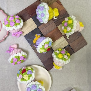 May include: A collection of handmade crochet flower bouquets in various colors, including pink, yellow, green, and purple. The bouquets are arranged on a wooden surface and a white plate, showcasing the delicate details of the yarn work.
